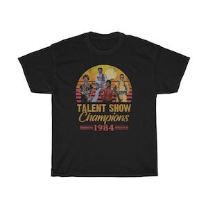 Op de afbeelding: Zwart T-shirt met een retro-grafiek van vier mensen in een band. De grafiek bevat een zonsondergang en de tekst "Talent Show Champions 1984".