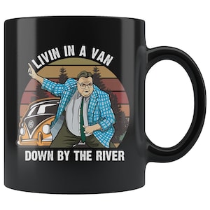 May include: Black coffee mug with a retro-style illustration of a man in a blue plaid jacket dancing in front of a yellow van with the text "Livin in a Van Down by the River".