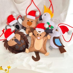 Puede incluir: Adornos de animales de fieltro, incluyendo un koala, un oso, un kiwi, una ardilla, una cacatúa y un pájaro. Cada adorno tiene un gorro de Papá Noel rojo y una cuerda roja para colgar. Los adornos son de color marrón, gris y blanco.