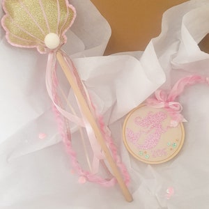 Puede incluir: Una varita de sirena rosa y dorada con una concha de mar de brillo y una cinta rosa. La varita est&aacute; en una bolsa de regalo de papel de seda blanco con un adorno de sirena rosa y blanco.