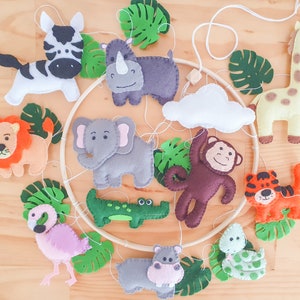 May include: A wooden mobile with felt animal and plant cutouts. The animals include a lion, elephant, rhino, giraffe, monkey, hippopotamus, crocodile, flamingo, and snake. The plants are green and leafy.