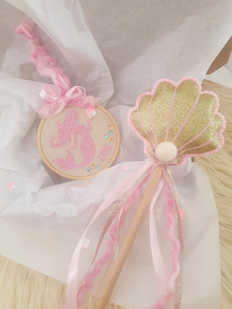 Puede incluir: Una varita de sirena rosa y dorada con una concha brillante y una silueta de sirena rosa en un aro. La varita tiene cintas rosas y blancas y un palo de madera.