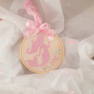 Puede incluir: Una silueta de sirena de purpurina rosa bordada en un aro de madera redondo con cinta rosa y una concha rosa. El aro est&aacute; rodeado de lentejuelas rosas e iridiscentes.