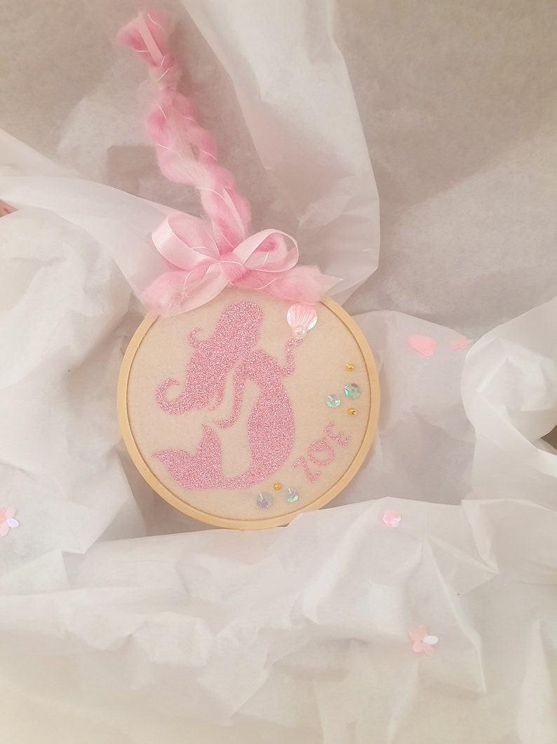 Puede incluir: Una silueta de sirena de purpurina rosa bordada en un aro de madera redondo con cinta rosa y una concha rosa. El aro est&aacute; rodeado de lentejuelas rosas e iridiscentes.