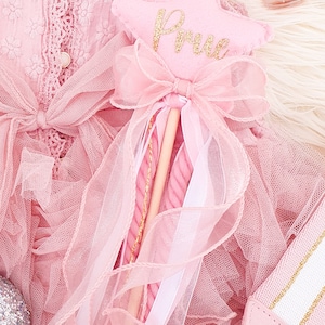 Puede incluir: Una varita de estrella de fieltro rosa con el nombre Prue en purpurina dorada, adornada con un lazo de organza rosa y cintas, sobre un vestido rosa texturizado con una correa a juego y un pequeño frasco de esmalte rosa.