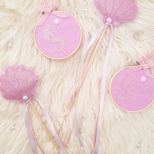 Puede incluir: Tres varitas de sirena rosa y dorado con detalles de purpurina y perlas. Las varitas tienen un palo de madera con cinta rosa y dorada. La parte superior de cada varita es un dise&ntilde;o diferente: una concha, una sirena con un globo y una sirena con un coraz&oacute;n. Las varitas est&aacute;n etiquetadas con "Amelia" y "Ellie".