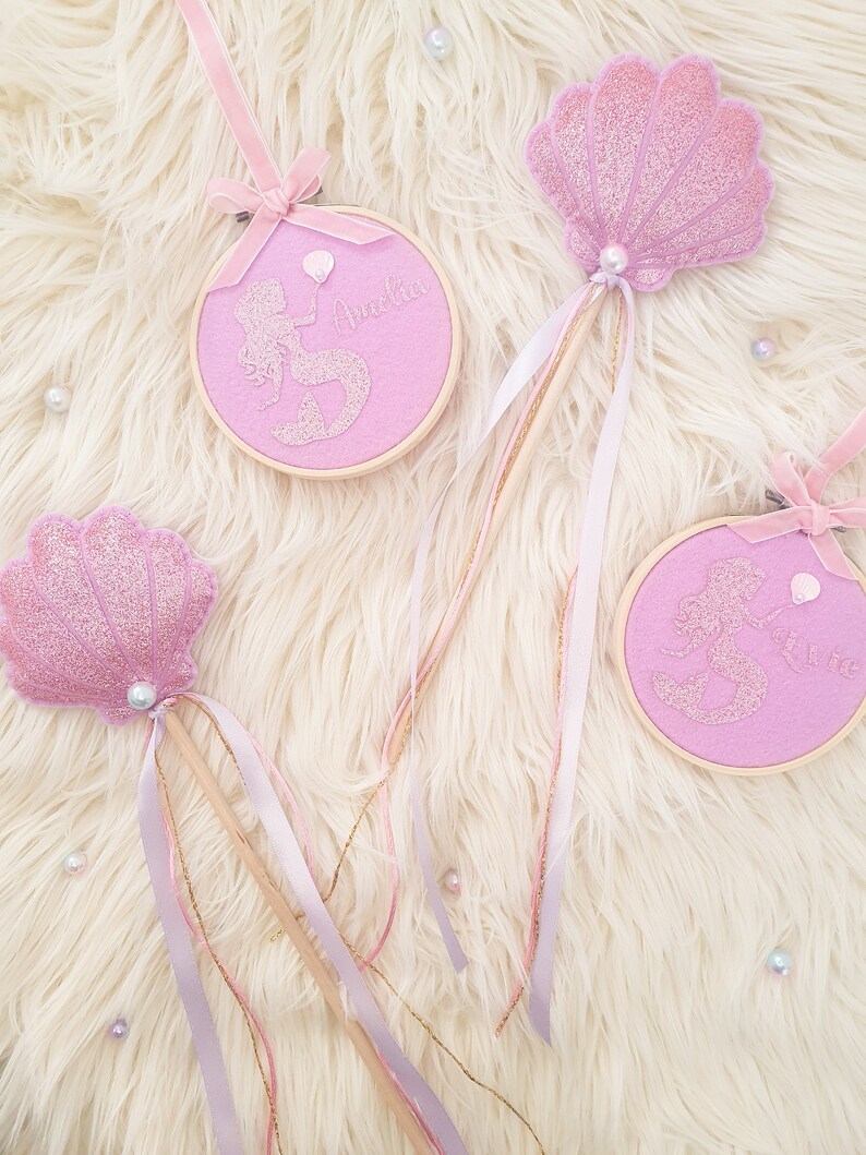 Puede incluir: Tres varitas de sirena rosa y dorado con detalles de purpurina y perlas. Las varitas tienen un palo de madera con cinta rosa y dorada. La parte superior de cada varita es un dise&ntilde;o diferente: una concha, una sirena con un globo y una sirena con un coraz&oacute;n. Las varitas est&aacute;n etiquetadas con "Amelia" y "Ellie".