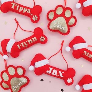 Puede incluir: Adornos navideños de fieltro rojo con forma de hueso y huella de perro, con detalles de purpurina dorada. Cada adorno de hueso tiene un nombre en escritura blanca y una pequeña huella de pata. Algunos tienen gorros de Papá Noel.
