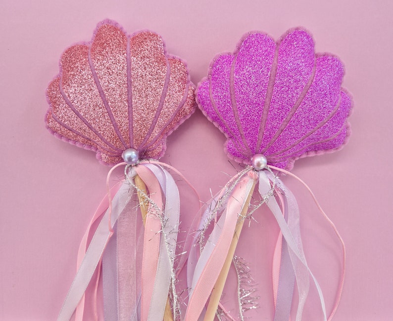 Puede incluir: Dos varitas de conchas de mar de purpurina rosa y p&uacute;rpura con cintas blancas y rosas. Las varitas son perfectas para una fiesta o un disfraz con tem&aacute;tica de sirenas.