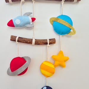 Puede incluir: Un móvil con temática espacial que presenta figuras de fieltro: un cohete azul claro, un planeta azul con anillo dorado, un planeta rojo con anillo plateado, una estrella amarilla, un planeta naranja a rayas y un planeta azul oscuro, colgados de dos ramas.