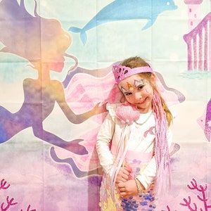 Puede incluir: Una ni&ntilde;a vestida de sirena con una cola de lentejuelas rosa, una camisa blanca y una corona rosa. Lleva una varita de concha rosa y extensiones de pelo moradas. El fondo es una pintura de acuarela de una sirena, un delf&iacute;n y un castillo.