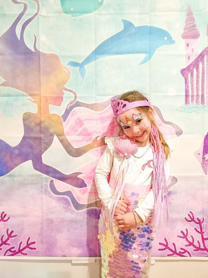 Puede incluir: Una ni&ntilde;a vestida de sirena con una cola de lentejuelas rosa, una camisa blanca y una corona rosa. Lleva una varita de concha rosa y extensiones de pelo moradas. El fondo es una pintura de acuarela de una sirena, un delf&iacute;n y un castillo.