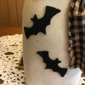 May include: A white fabric jar with two black felt bat cutouts. The jar is adorned with a black and tan plaid fabric scarf. The jar sits on a white lace doily, creating a Halloween theme.