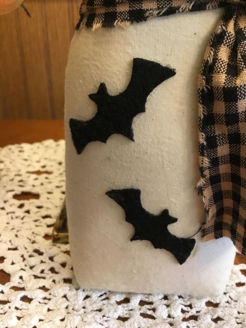 May include: A white fabric jar with two black felt bat cutouts. The jar is adorned with a black and tan plaid fabric scarf. The jar sits on a white lace doily, creating a Halloween theme.
