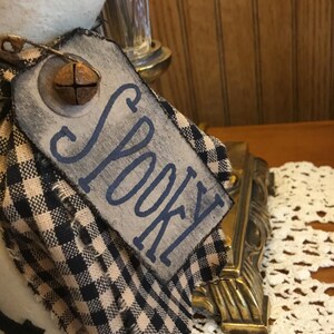May include: A decorative tag with the word "Spooky" in blue lettering. The tag is attached to a black and tan checkered fabric. A small, rusty bell is attached to the tag. The tag is part of a Halloween-themed decoration.
