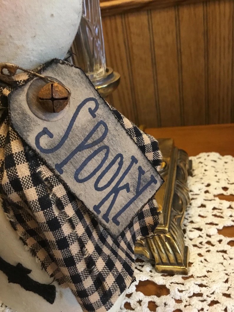 May include: A decorative tag with the word "Spooky" in blue lettering. The tag is attached to a black and tan checkered fabric. A small, rusty bell is attached to the tag. The tag is part of a Halloween-themed decoration.
