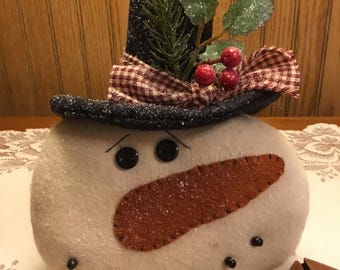 May include: A snowman decoration with a black top hat, a brown carrot nose, and black button eyes. The hat is decorated with a plaid ribbon, holly leaves, and red berries. The snowman is made of a soft, white material and has black button eyes and a stitched mouth.