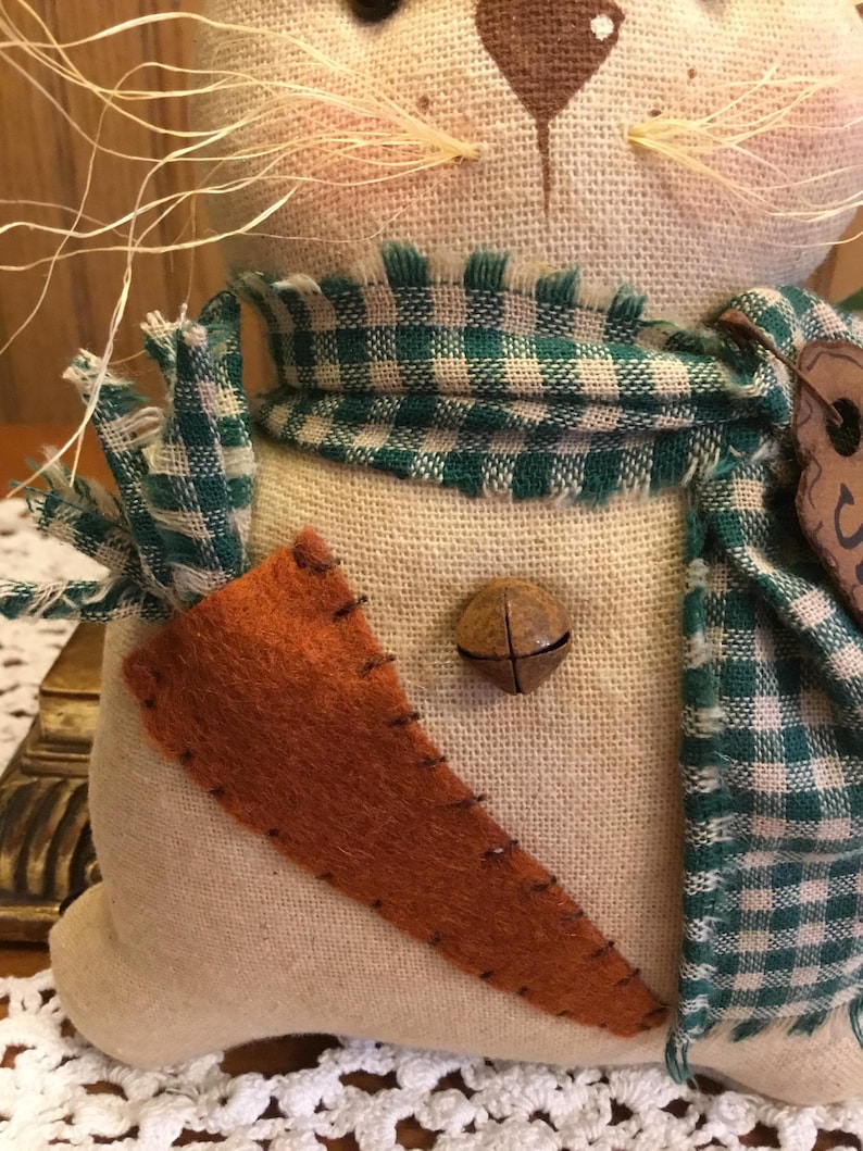 May include: A handmade plush bunny toy with a green and white checkered scarf, a brown felt carrot patch, and a brown bell. The bunny has a beige fabric body and a brown nose.