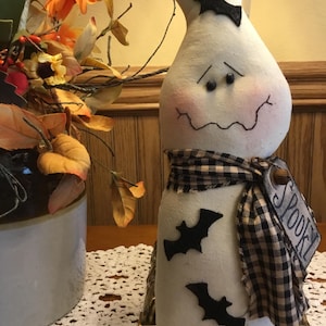 May include: A white Halloween ghost decoration with a curved head, black eyes, and a stitched mouth. It has a black and white checkered scarf and black bat cutouts. A tag reads "Spooky".