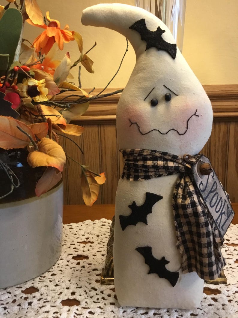 May include: A white Halloween ghost decoration with a curved head, black eyes, and a stitched mouth. It has a black and white checkered scarf and black bat cutouts. A tag reads "Spooky".