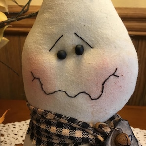 May include: A handmade, pear-shaped ghost doll with a black bow, black button eyes, and a stitched mouth. It wears a black and tan checkered scarf and a tag with the word "Home".