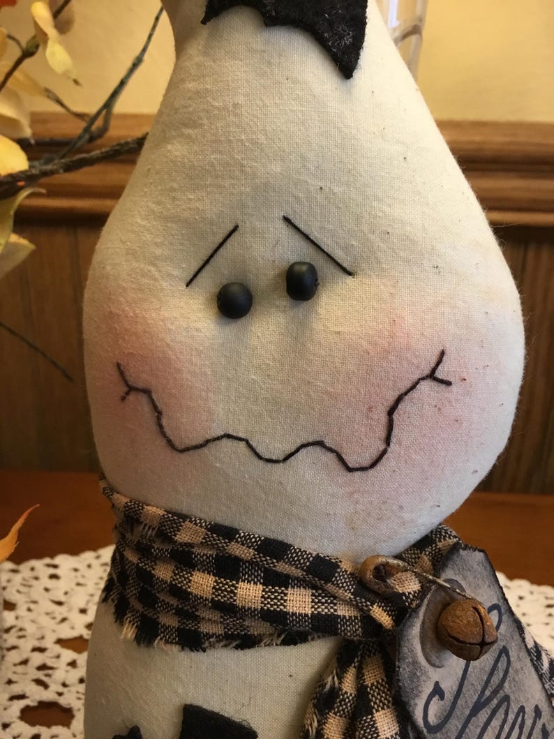 May include: A handmade, pear-shaped ghost doll with a black bow, black button eyes, and a stitched mouth. It wears a black and tan checkered scarf and a tag with the word "Home".