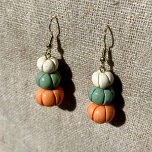 Könnte beinhalten: Ohrringe mit einem gestapelten Kürbis-Design. Jeder Ohrring hat drei Kürbisse in Weiß, Grün und Orange, mit goldfarbenen Ohrhaken. Die Ohrringe sind vor einem neutralen Hintergrund platziert.