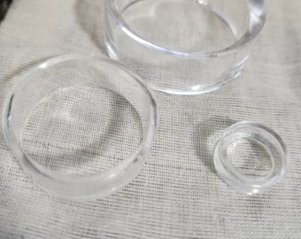 Puede incluir: Tres anillos transparentes de acrílico de diferentes tamaños se muestran sobre una superficie clara y texturizada. Los anillos son transparentes, lo que permite ver el fondo a través de ellos. El anillo más grande tiene un diámetro de unos 10 cm, el más pequeño, unos 2,5 cm.