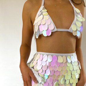 May include: White iridescent sequin top and skirt set. The top is a halter style with a triangle shape. The skirt is a mini skirt with a high waist.