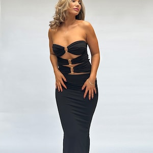 May include: A black strapless maxi dress with gold ring detailing. The dress has a fitted bodice and a flowing skirt.