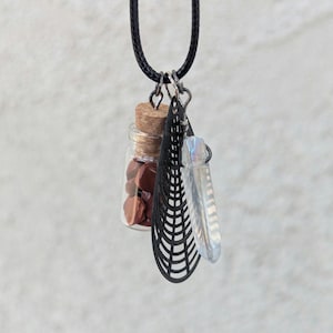 May include: A pendant necklace featuring a small glass bottle filled with red stones, a black filigree charm, and a clear crystal. The necklace has a black cord and silver-tone metal accents. The bottle is sealed with a cork.