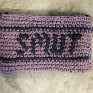 May include: A handmade crochet pouch in shades of mauve and charcoal gray. The word "SPARK" is prominently displayed in charcoal gray across the center of the pouch. The pouch is rectangular and appears to be made of soft yarn.