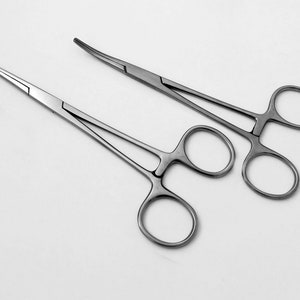 Puede incluir: Dos hemostáticos de acero inoxidable, también conocidos como pinzas mosquitos, están sobre una superficie blanca. Los hemostáticos están cerrados y tienen una punta curva.