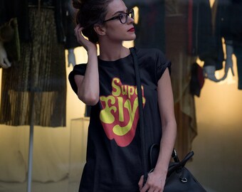 May include: Black t-shirt with the text "Super Fly" in yellow and red. The shirt is worn with denim shorts and black over-the-knee socks. A black purse is worn over the shoulder.