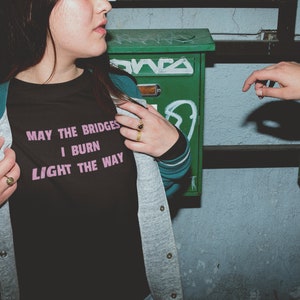 Può includere: Una persona indossa una maglietta nera con il testo "May the bridges I burn light the way" in lettere rosa. La persona indossa una giacca grigia sopra la maglietta e ha un anello d'oro al dito.