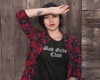 May include: A person wearing a black t-shirt with the text "Bad Girls Club" in white lettering. The person is wearing a red and black plaid shirt over the t-shirt.