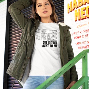Könnte beinhalten: Ein weißes T-Shirt mit schwarzem Text, der "Oh Sit Down Oh Sit Down Oh Sit Down Sit Down Next To Me" lautet. Die Person, die das Shirt trägt, steht auf einer grünen Treppe.