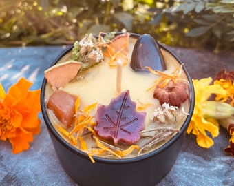 May include: A black candle jar with a yellow wax base and a white wick. The candle is decorated with dried flowers, leaves, and small stones. The jar is sitting on a gray surface.