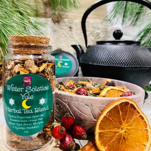 Puede incluir: Un frasco de vidrio lleno de una mezcla de hierbas y especias secas para una infusión de hierbas Yule de solsticio de invierno. La etiqueta dice "Winter Solstice Yule Herbal Tea Blend" y enumera los ingredientes: Rooibos, Menta piperita, Menta verde, Corteza de canela, Cáscara de naranja, Raíz de jengibre, Clavo de olor, Pimienta de Jamaica, Escaramujos, Cardamomo. El frasco está sobre una superficie de madera con un tazón de frutas secas y especias, una rama de canela y algunas bayas rojas.