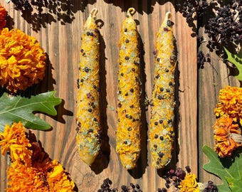 Può includere: Tre candele gialle decorate con erbe e bacche essiccate sono disposte su una superficie di legno, circondate da fiori di calendula arancioni, foglie verdi e bacche scure. Le candele hanno un'estetica rustica e naturale.