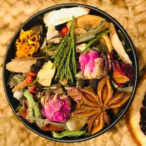 May include: A close-up of a small, round tin filled with a variety of dried herbs, spices, and flowers. The tin is filled with a mix of brown, green, yellow, and pink ingredients.