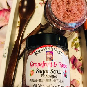 May include: A glass jar of pink grapefruit and rose sugar scrub with a black lid. The label reads "NEW DAWN ELEMENTS Grapefruit & Rose Sugar Scrub Hancrafted ENERGIZE MOISTURIZE INVIGORATE All Natural Skin Care Nt Wt. 4 oz".