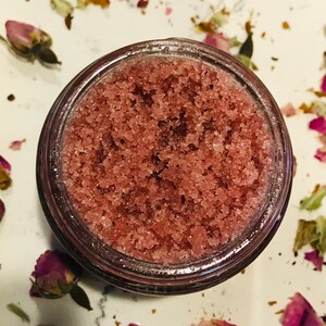 May include: A close-up of a glass jar filled with pink sugar scrub. The scrub is a mixture of sugar and a pink liquid, creating a grainy texture. The jar is sitting on a white surface with dried rose petals scattered around it.