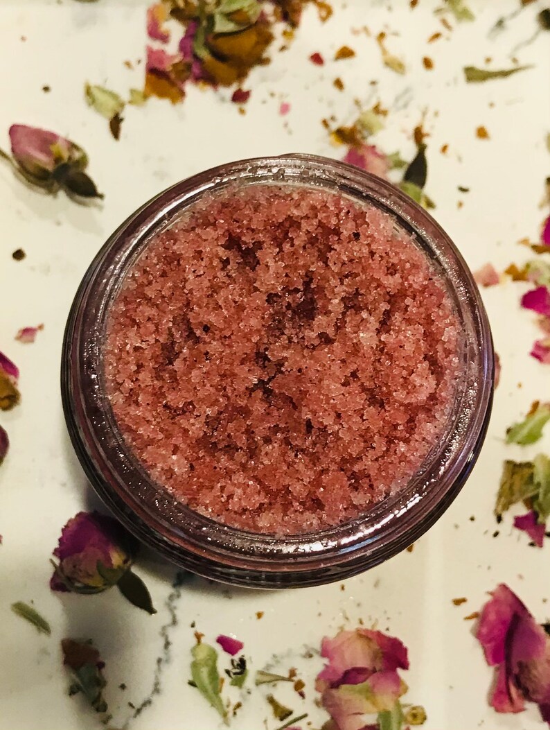 May include: A close-up of a glass jar filled with pink sugar scrub. The scrub is a mixture of sugar and a pink liquid, creating a grainy texture. The jar is sitting on a white surface with dried rose petals scattered around it.