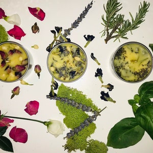 Op de afbeelding: Drie kleine kaarsen in metalen blikken met verschillende botanische elementen ingebed in de was. De kaarsen zijn omringd door gedroogde bloemen, kruiden en bladeren, waaronder lavendel, rozemarijn en basilicum.