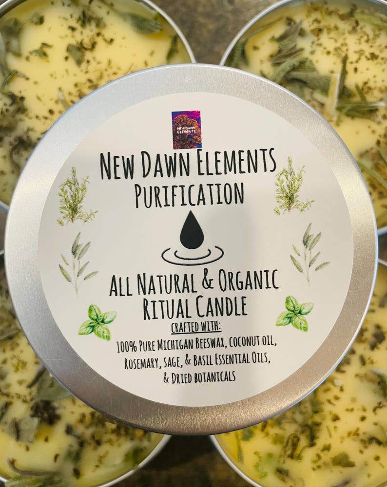 Op de afbeelding: Een zilveren blikje met een kaars met een gele wax bovenkant en gedroogde kruiden. Het etiket luidt "New Dawn Elements Purification All Natural & Organic Ritual Candle Crafted With: 100% Pure Michigan Beeswax, Coconut Oil, Rosemary, Sage, & Basil Essential Oils, & Dried Botanicals"
