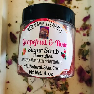 May include: A glass jar of grapefruit and rose sugar scrub with a black lid. The label reads "New Dawn Elements Grapefruit & Rose Sugar Scrub Hancrafted Energize Moisturize Invigorate All Natural Skin Care Nt Wt. 4 oz".