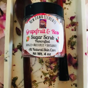 May include: A glass jar with a black lid filled with a pink and white sugar scrub. The label reads "New Dawn Elements Grapefruit & Rose Sugar Scrub Hancrafted Energize Moisturize Invigorate All Natural Skin Care Nt Wt. 4 oz".
