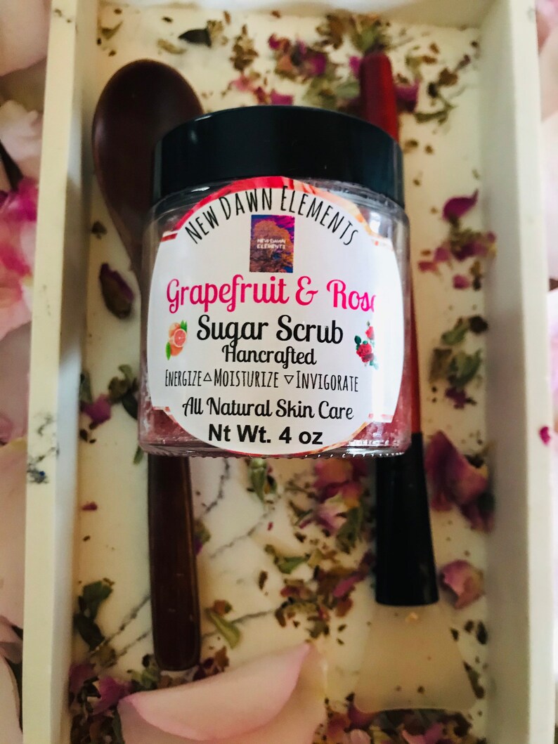 May include: A glass jar with a black lid filled with a pink and white sugar scrub. The label reads "New Dawn Elements Grapefruit & Rose Sugar Scrub Hancrafted Energize Moisturize Invigorate All Natural Skin Care Nt Wt. 4 oz".