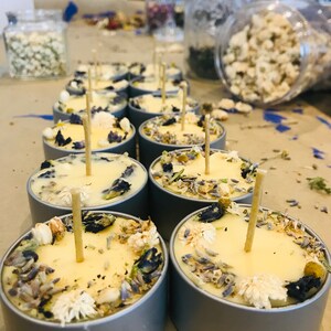 Op de afbeelding: Een close-up van een groep kleine, ronde, gele kaarsen met gedroogde bloemen en kruiden die in de was zijn ingebed. De kaarsen bevinden zich in zilveren containers en hebben houten lonten.